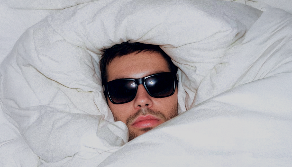 A man wearing sunglasses wrapped in bedding so that only his face is visible