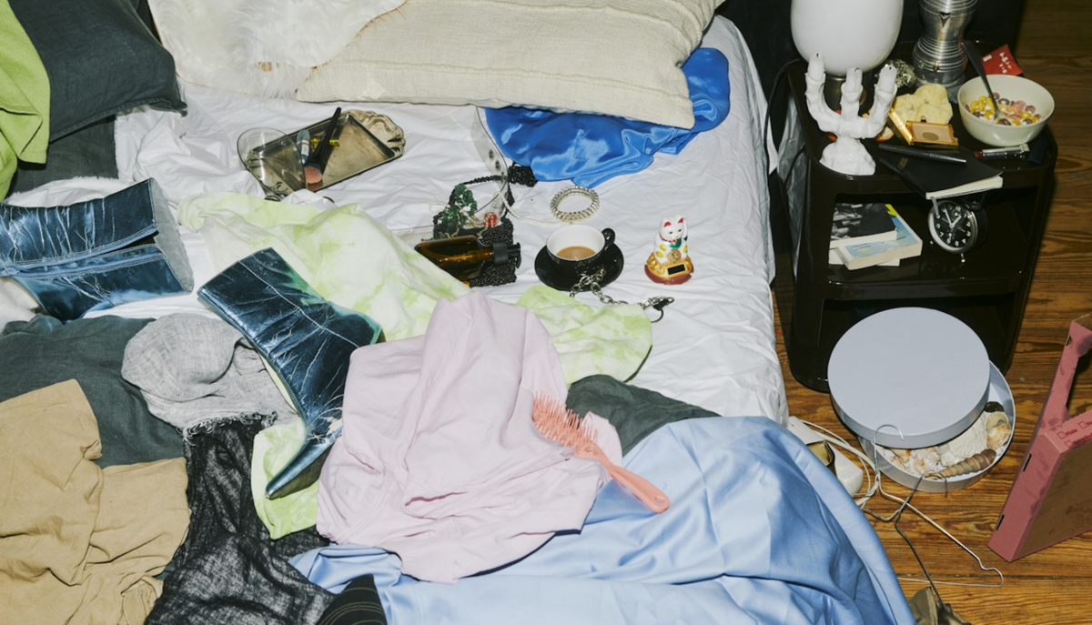 A messy, unmade bed covered in colourful clothes and boots, a coffee cup, and everyday objects.